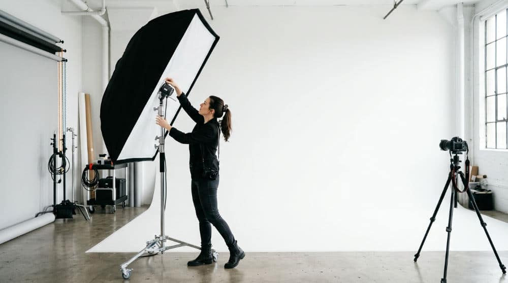 A professional photographer setting up studio lighting in front of a white cyclorama wall for a headshot.