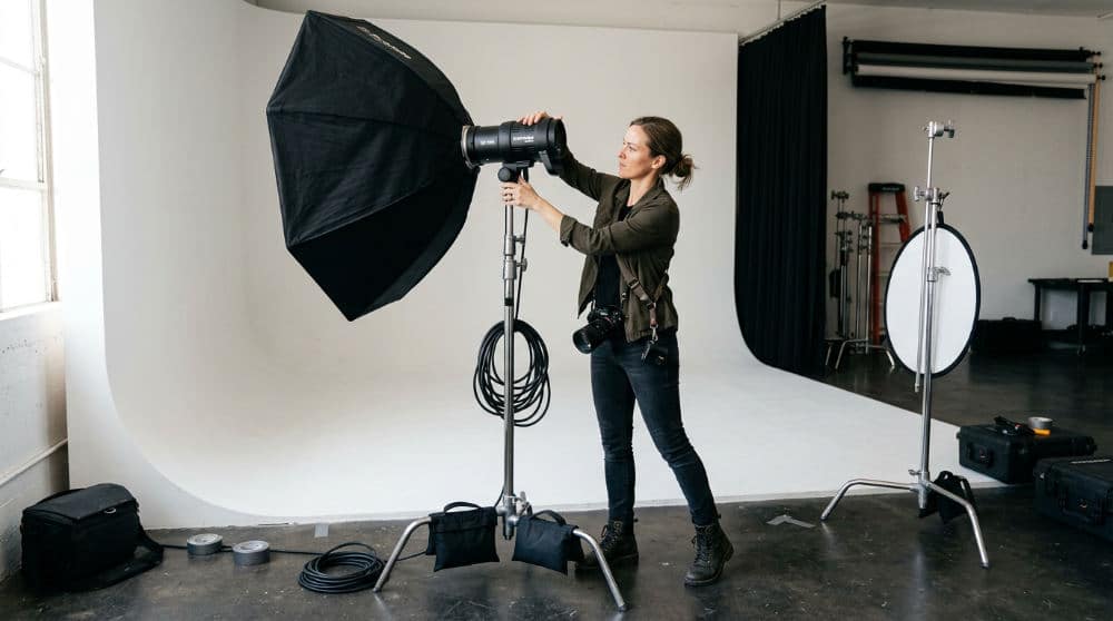 A photographer setting up high-output studio lighting next to a large white photography cyclorama wall.