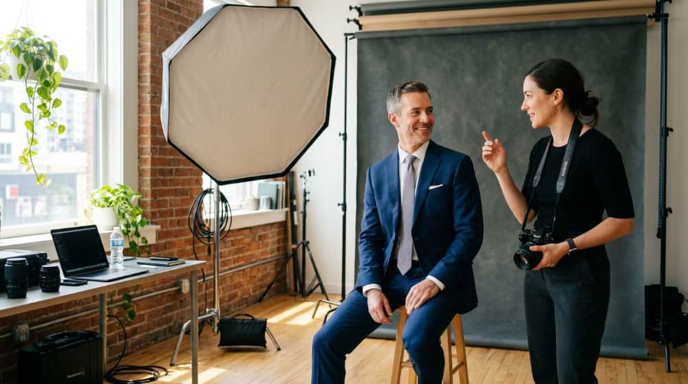 A male photographer coaching a professional woman during a headshot session in front of a neutral backdrop.